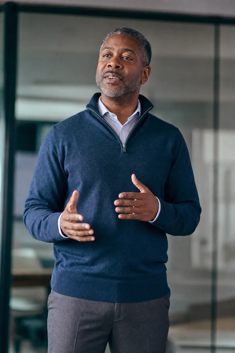 A man in a navy zip sweater briefs his team to camera from a glass-walled office.