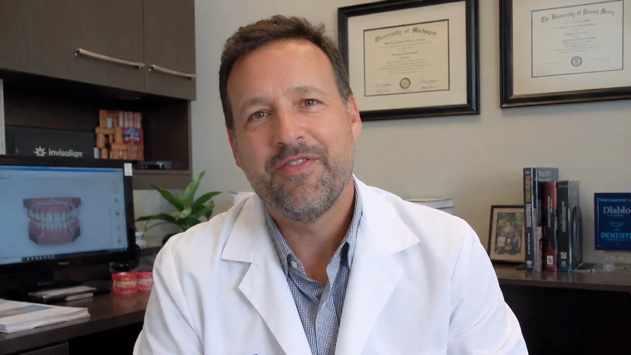 An orthodontist in a white coat records a welcome in front of his framed diploma.